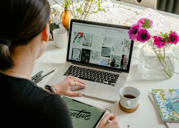Woman in a creative workspace using a laptop and tablet for calligraphy. Artistic and tech-driven environment.